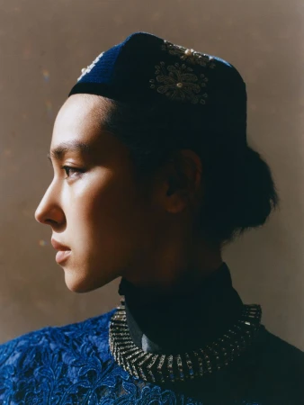 Profile portrait of a woman wearing an embroidered dark blue cap and matching textured garment with a statement necklace.