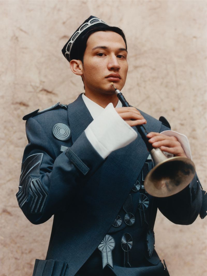 Portrait of a man playing a wind instrument, wearing a decorated blue jacket and hat against a mottled light brown backdrop.