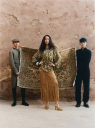 Three models posing against an earth wall, a woman centrally flanked by two men holding a patterned rug