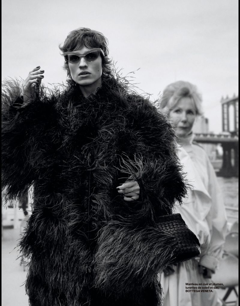 A monochrome fashion shot of a woman in sunglasses and a black feather coat, walking outdoors with another woman in the background.