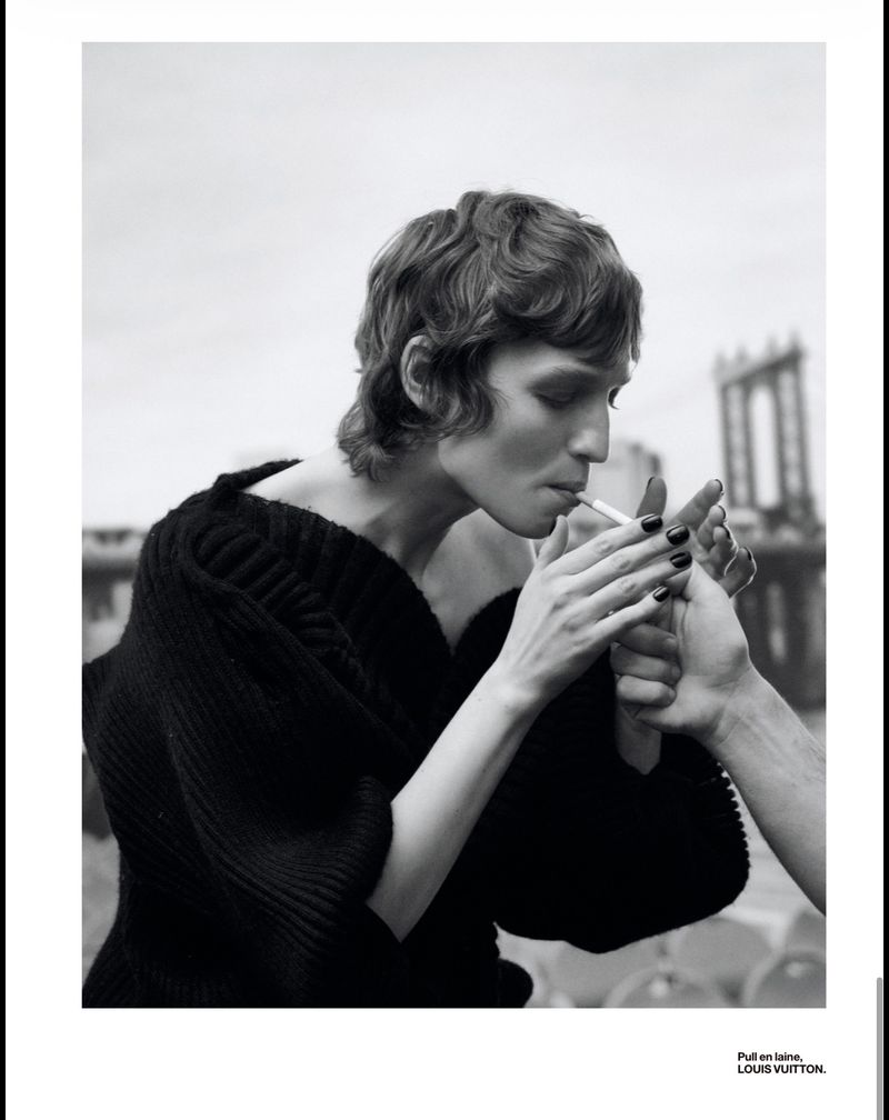 Monochrome fashion portrait of a woman in a dark sweater smoking a cigarette, lit by another person, with a bridge backdrop.