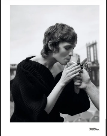 Monochrome fashion portrait of a woman in a dark sweater smoking a cigarette, lit by another person, with a bridge backdrop.