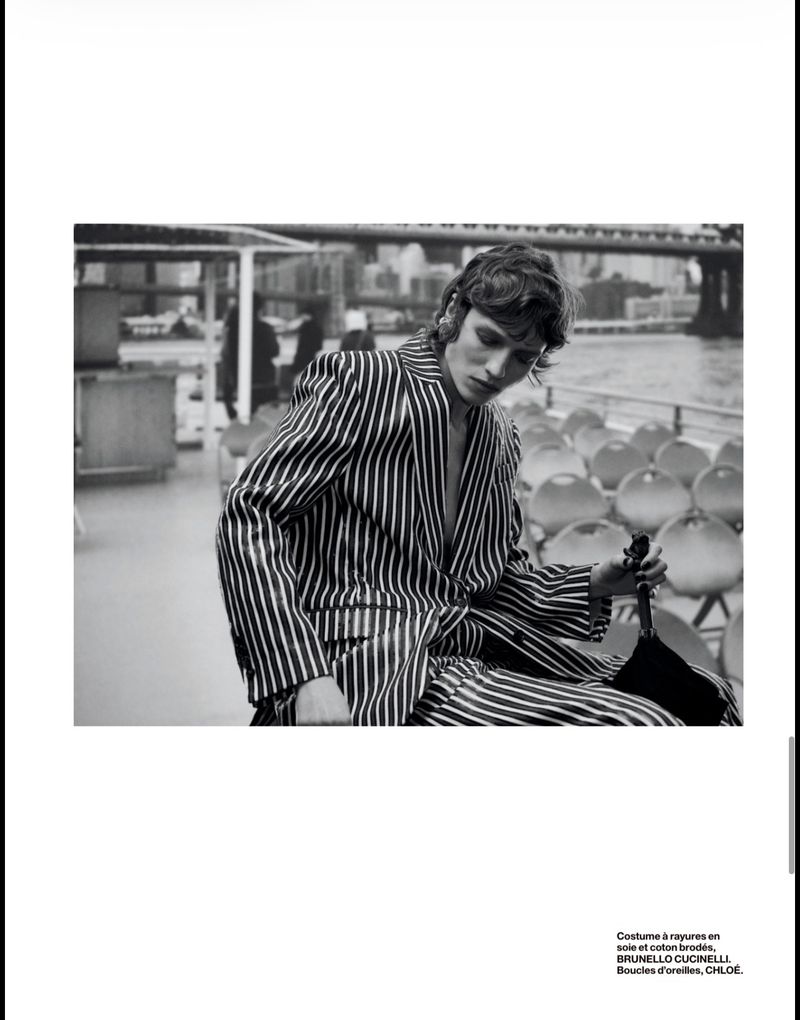 Black and white fashion photograph featuring a male model in a striped suit sitting on a boat on a river with a bridge in the background.