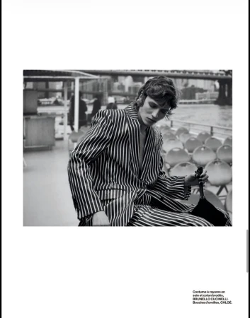 Black and white fashion photograph featuring a male model in a striped suit sitting on a boat on a river with a bridge in the background.
