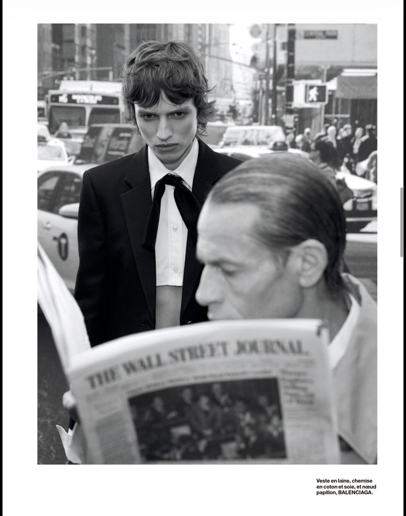 Black and white fashion shot with a young adult male in a suit standing behind an older man reading The Wall Street Journal in a city.