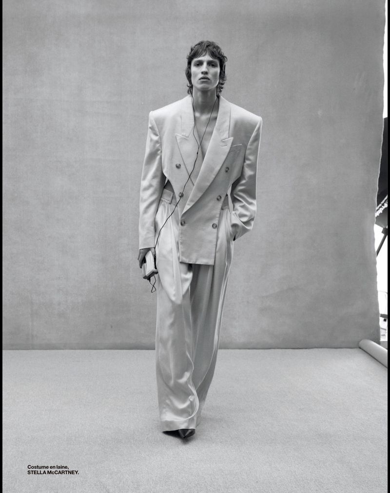 Fashion shot of a slender adult male with brown hair wearing an oversized, double-breasted suit with wide-leg trousers in front of gray backdrop.