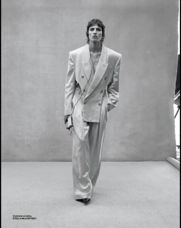 Fashion shot of a slender adult male with brown hair wearing an oversized, double-breasted suit with wide-leg trousers in front of gray backdrop.