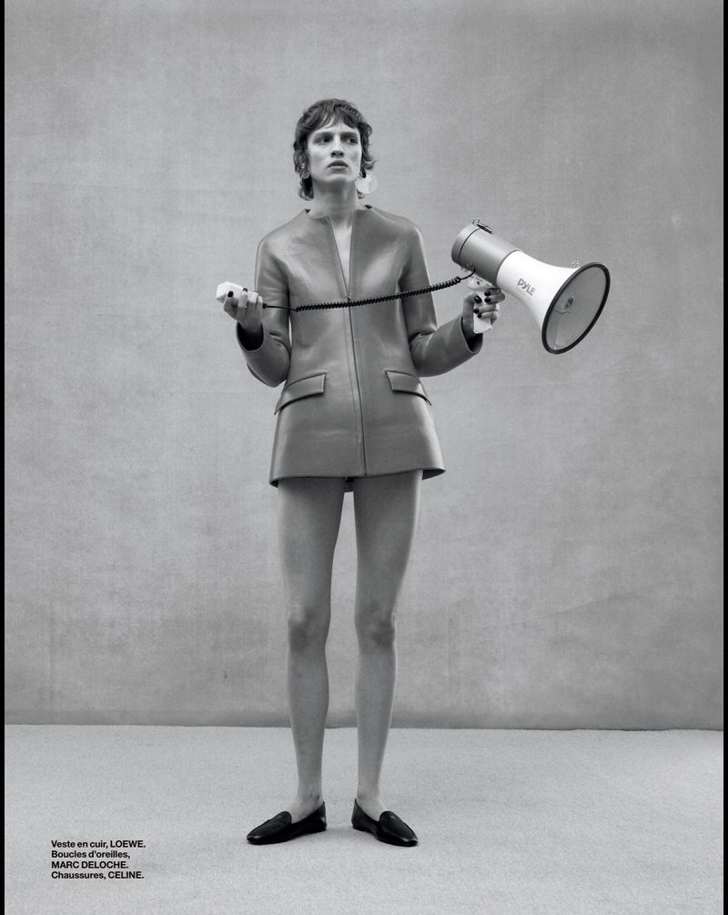 A slender adult female stands in a photo studio wearing a brown leather jacket and black loafers while holding a megaphone and trigger.
