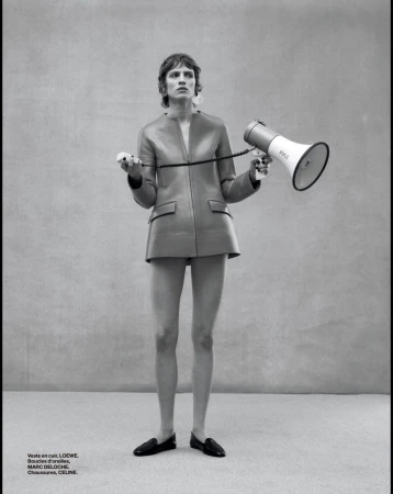A slender adult female stands in a photo studio wearing a brown leather jacket and black loafers while holding a megaphone and trigger.