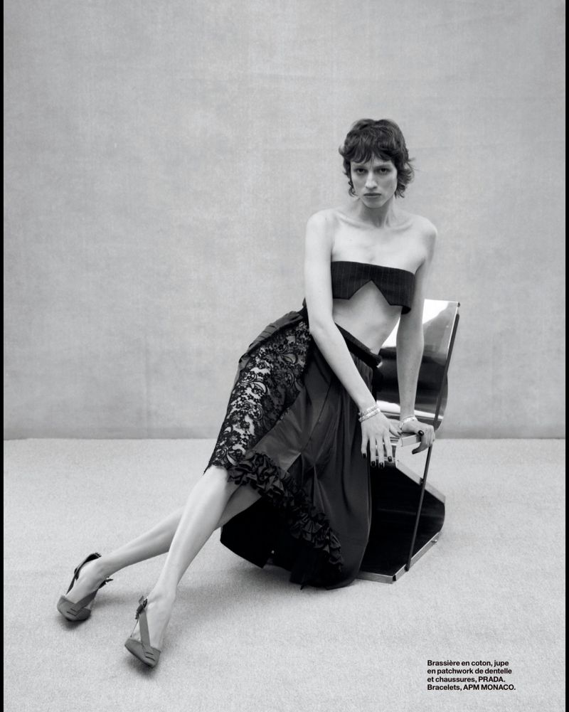 A woman is sitting on a chair wearing a bralette, a skirt with lace details, and slingback heels against a neutral-toned background.