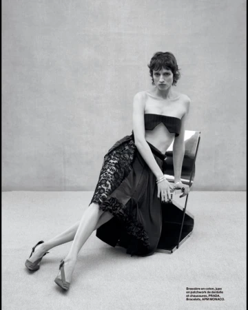 A woman is sitting on a chair wearing a bralette, a skirt with lace details, and slingback heels against a neutral-toned background.