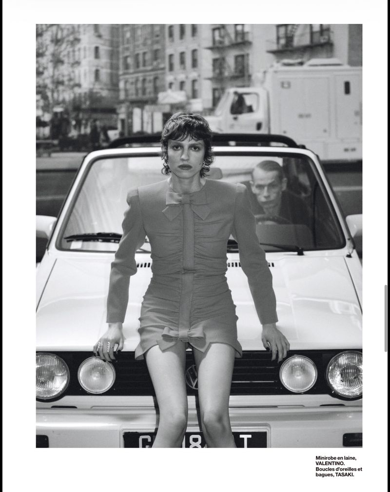Monochrome fashion editorial portrait of a woman in a dress leaning on a car in an urban setting, with a man visible in the back.