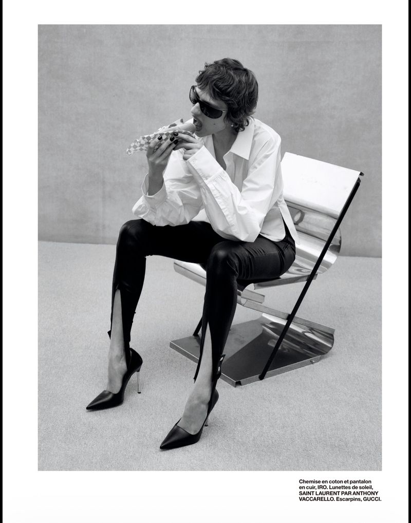 Model wearing dark sunglasses, white shirt, and black leather pants sitting on a metal chair eating food