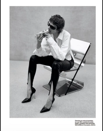 Model wearing dark sunglasses, white shirt, and black leather pants sitting on a metal chair eating food