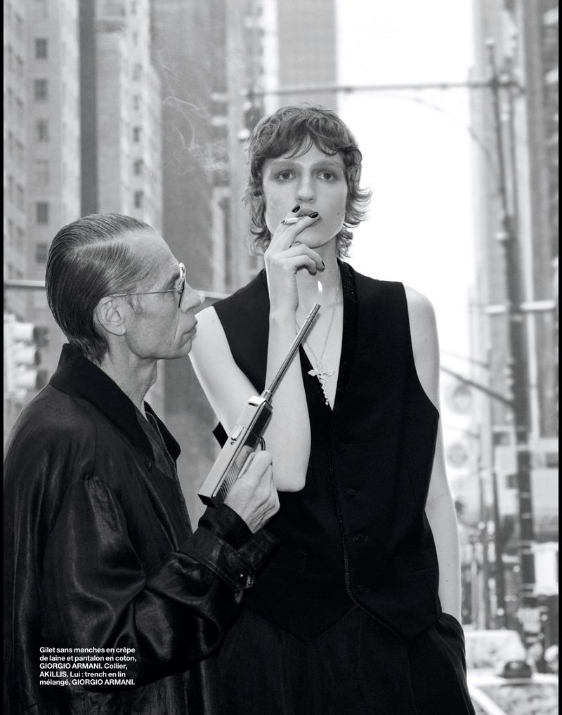 Fashion shot in black and white featuring two figures against city backdrop. One holds a gun, the other smokes with buildings behind.