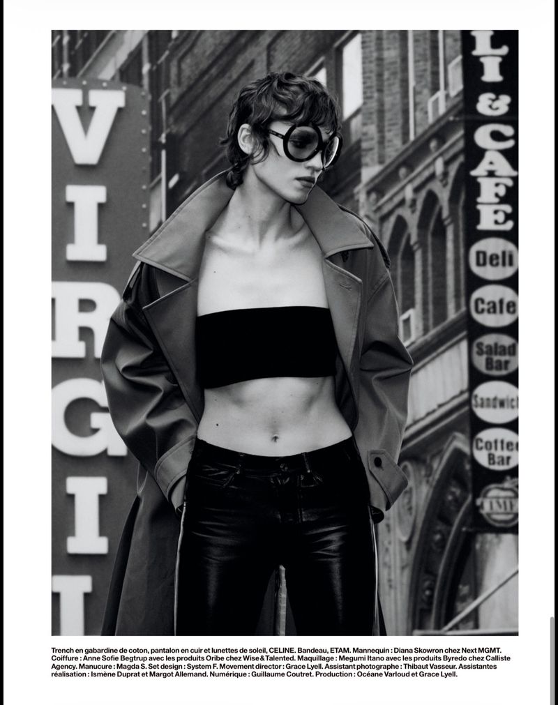 A young adult female fashion model poses in an urban outdoor setting, wearing an open trench coat, bandeau top, sunglasses, and leather trousers.