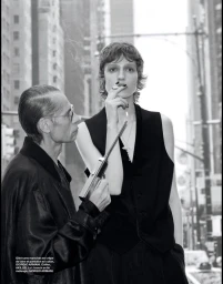Model in black vest smoking a cigarette while standing beside an older man holding a lighter on a city street.