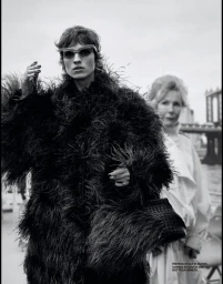 Model in black feathered coat wearing sunglasses outdoors with another person standing in the background.