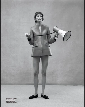 Model standing on a grey seamless studio background holding a megaphone and a small controller, wearing a jacket and black loafers.