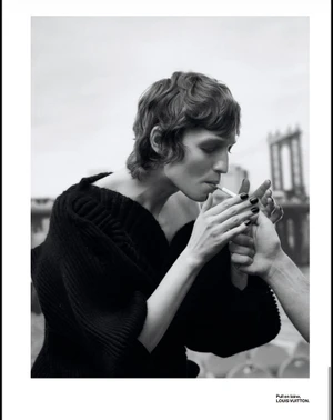 Model with short wavy hair wearing a black knitted sweater lighting a cigarette outdoors with a bridge visible