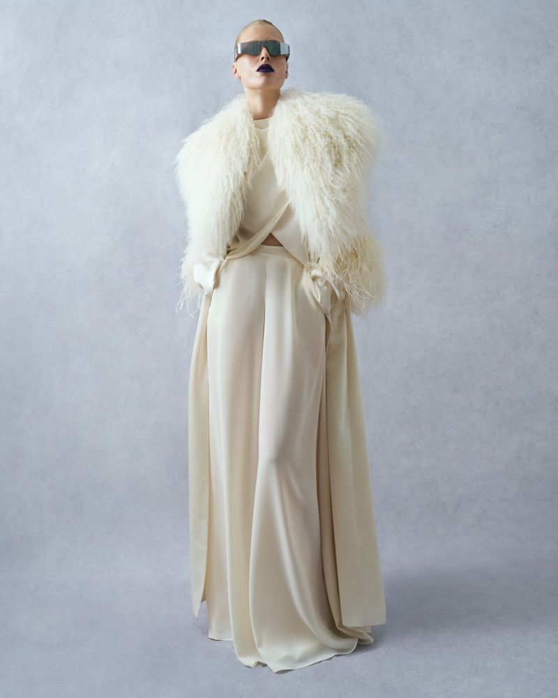 Adult fashion model poses in an off-white outfit with fluffy feather accents and dark lipstick against a textured grey backdrop.