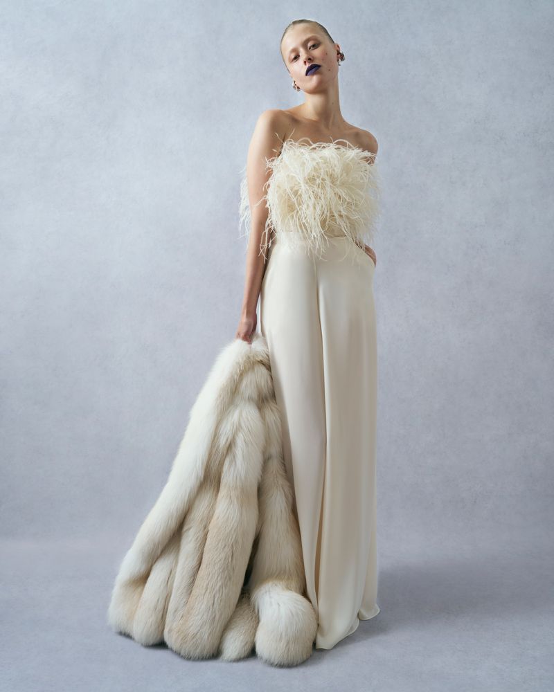 A woman in a feathered top and white pants holds a large fur coat in a studio setting, posing confidently with dark lipstick.