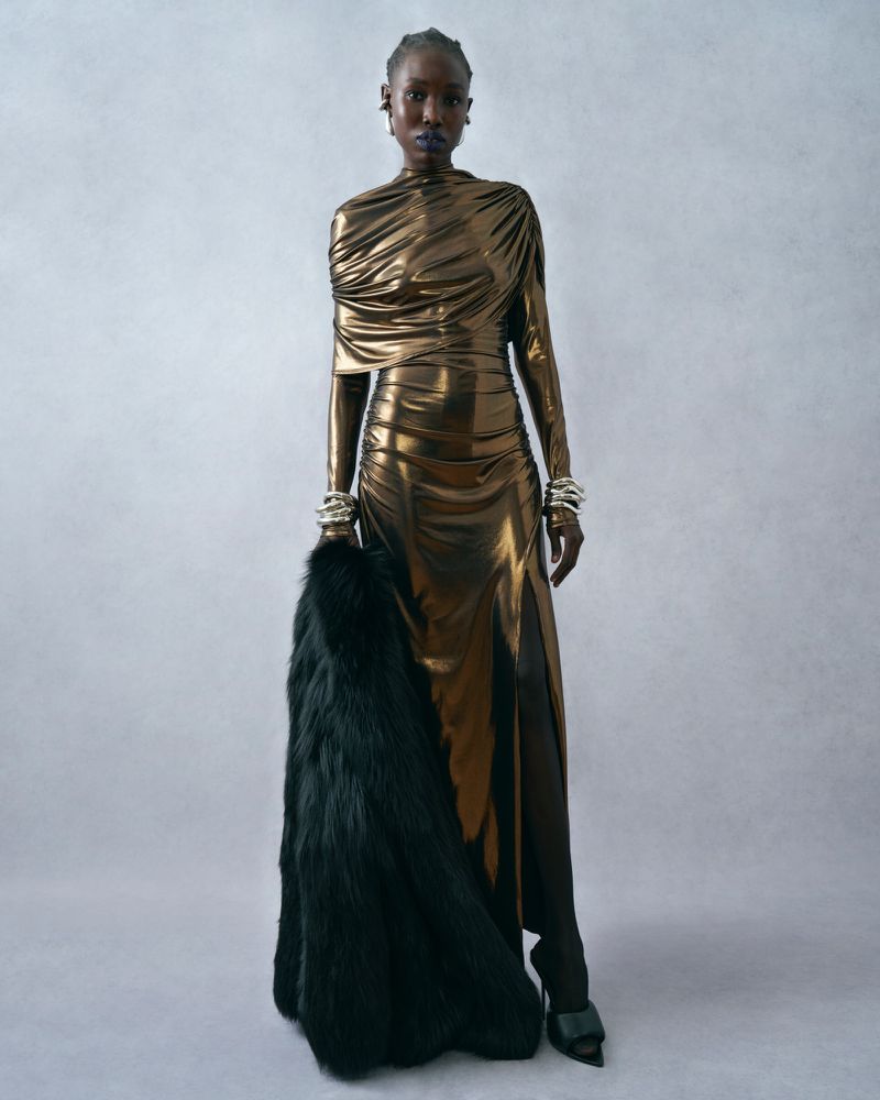A Black woman in a metallic gold gown and black heels poses with a fur coat in an indoor studio environment for a fashion editorial.