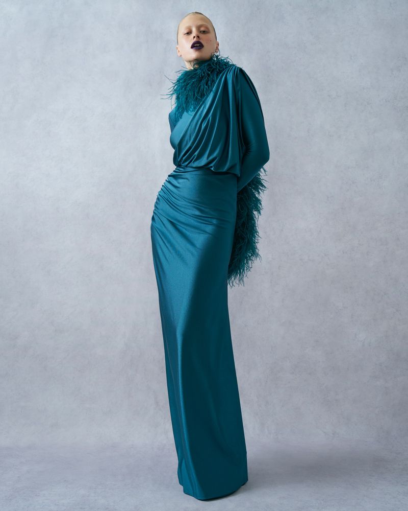 Adult female fashion model posing in a long teal gown with a feathered trim against a soft gray background in a studio setting.