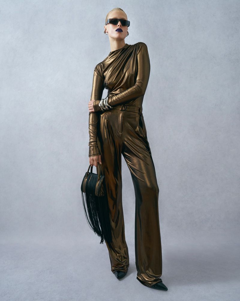 Fashionable adult female model in gold metallic-look outfit with sunglasses holding a fringe purse, posing in a photo studio.