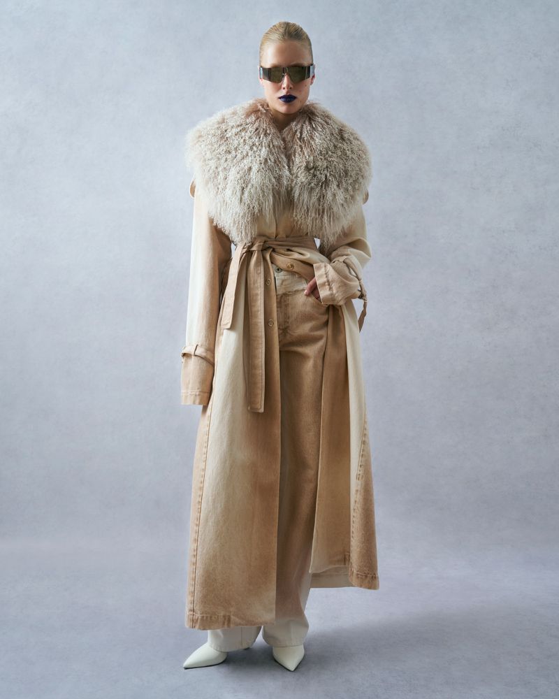 A woman poses in a long trench coat with fur collar, sunglasses, and white shoes in a studio setting with neutral background.
