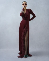Model in a long burgundy gown with a feather-trimmed thigh-high slit and dark sunglasses against a plain grey textured backdrop.