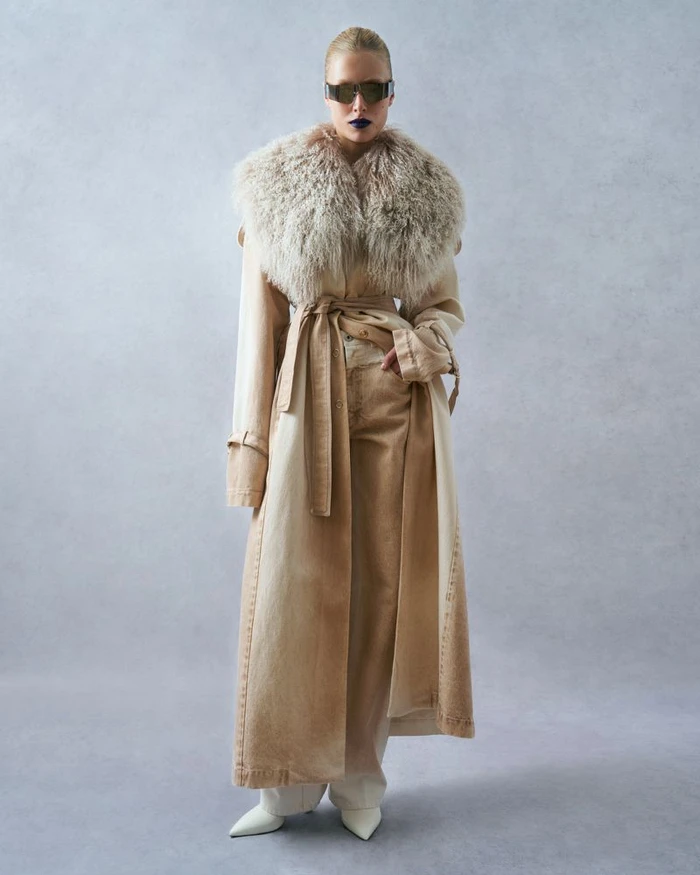 Model wearing a long beige belted coat with a furry collar, sunglasses, and matching trousers against a grey seamless background