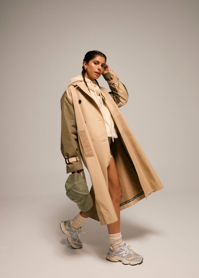 A woman wearing a beige trench coat with a beige hoodie and athletic shoes, holding a green baseball cap in a studio setting.