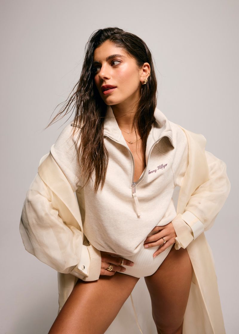 Brunette woman in studio, wearing Tommy Hilfiger top and coat, with shiny legs, posing against a neutral gray background.