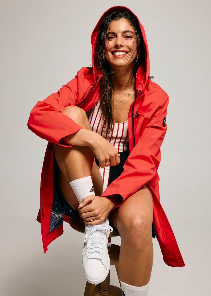 A smiling woman with dark hair in a red hooded raincoat sitting on a stool with white socks and tennis shoes in a photo studio.