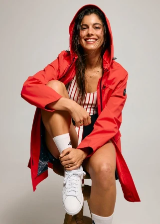 Smiling woman seated on a stool wearing a red hooded raincoat over a striped top and white sneakers against a grey background.