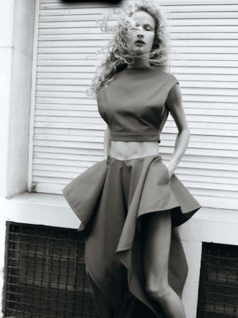 Fashion shot of a woman in a two-piece garment with a high-waisted skirt posing in front of a white-painted shuttered window.