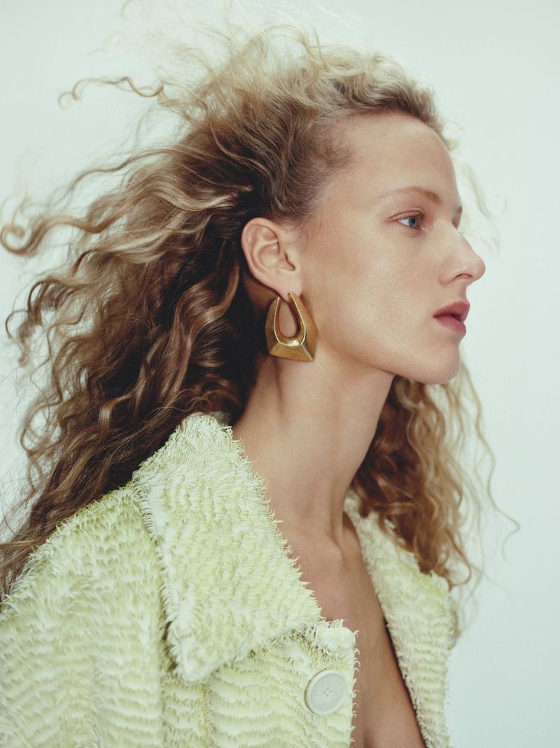 Profile portrait of a blonde woman wearing a lime green jacket and gold earrings against a light background in a beauty shot.