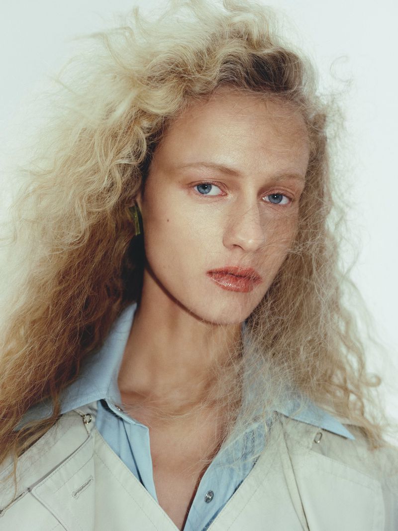 Close-up portrait of a fair-skinned woman with flowing blonde hair, wearing a light-blue collared shirt and neutral jacket against a white background.