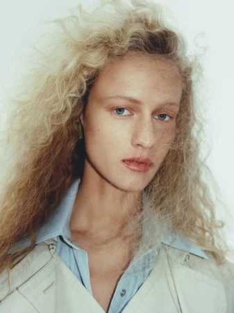Close-up portrait of a fair-skinned woman with flowing blonde hair, wearing a light-blue collared shirt and neutral jacket against a white background.