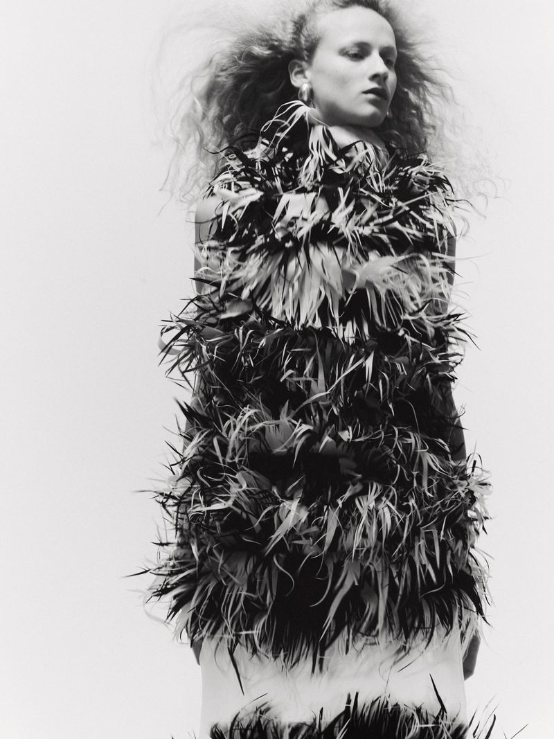 Monochrome portrait of a woman with fair skin and voluminous blonde hair wearing a tiered feather dress, captured in a studio setting.