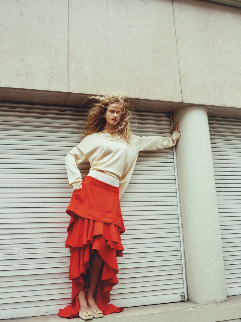 A woman with curly hair poses in a ruffled skirt and cream top against a textured wall, looking at the camera in an urban setting.
