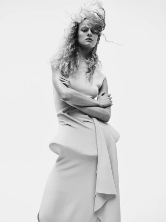 Model with curly hair and crossed arms wearing a sculpted dress against a plain white background