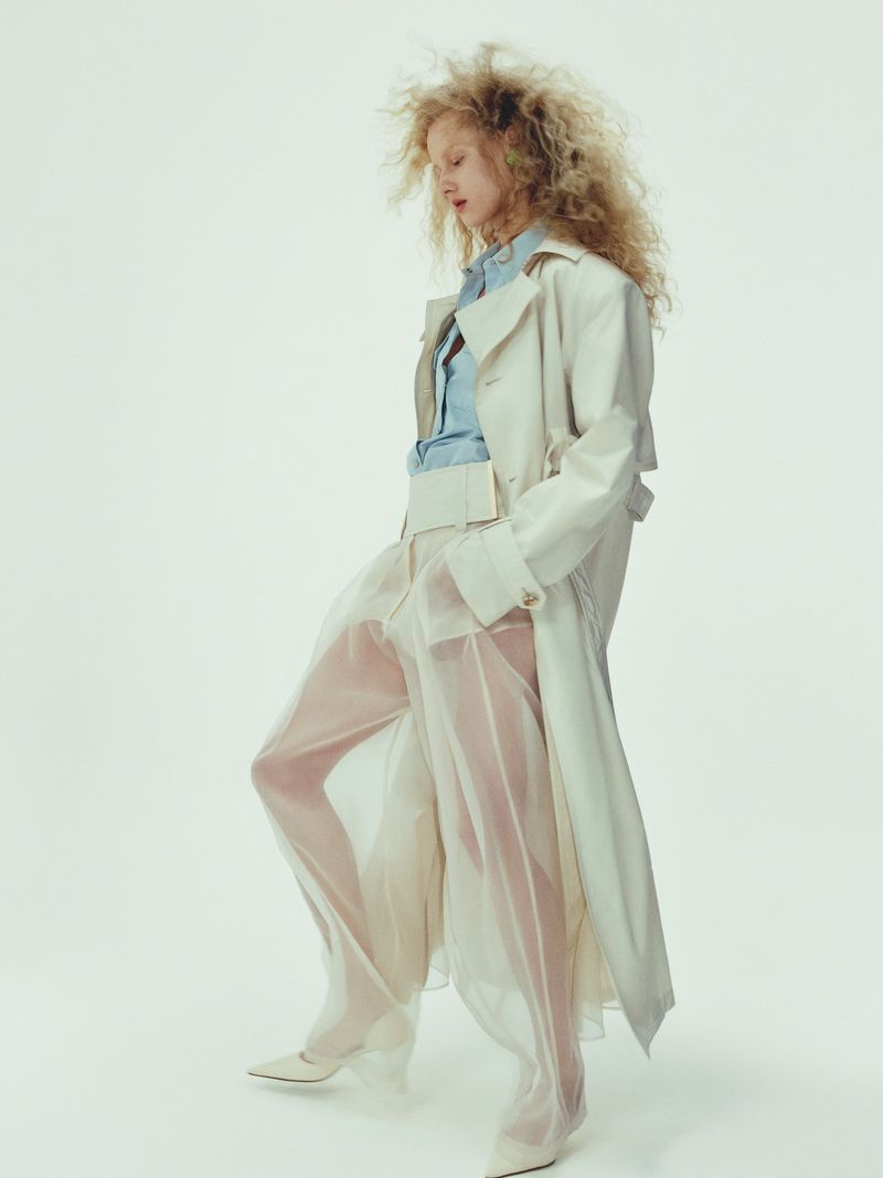 A fair-skinned woman with curly blonde hair poses in a long, light trench coat with semi-sheer pants and pointed-toe shoes in a studio.