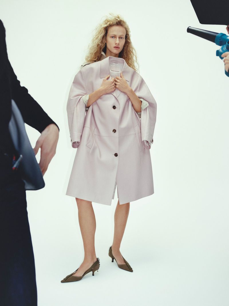 A young adult female poses in a pale pink trench coat and pumps against a white backdrop in a fashion studio with crew members visible.