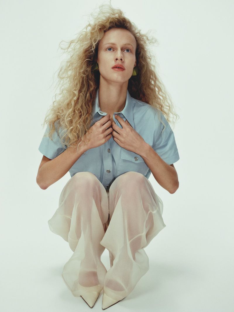 A woman with long, curly blonde hair and blue shirt is squatting while posing in a photo studio against a plain white background.