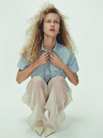 A woman with long, curly blonde hair and blue shirt is squatting while posing in a photo studio against a plain white background.