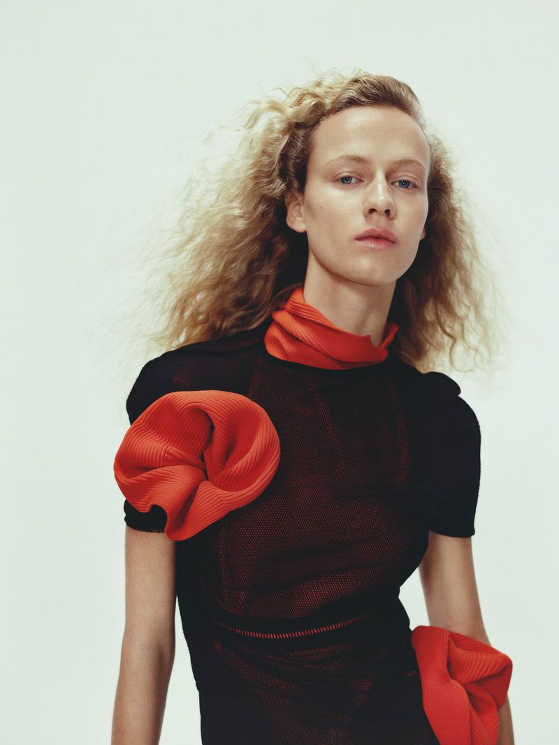 Woman with fair skin and curly blonde hair wears a sheer black top with orange, gathered fabric accents against a white backdrop.