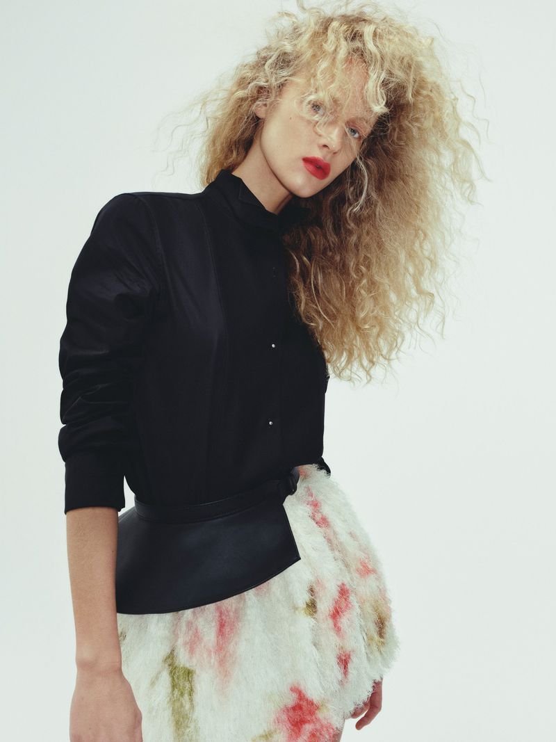 A curly-haired blonde model poses in a black top and patterned skirt against a white background in a fashion-style medium shot.