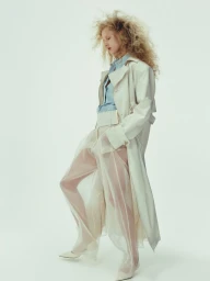 Model with voluminous blonde curly hair wearing a light trench coat, blue button-down shirt, and sheer wide-leg trousers against a white backdrop.